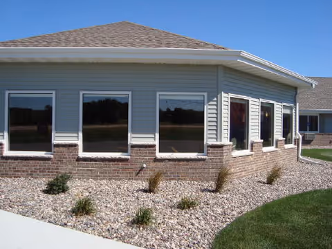 Exterior view of a single-story building with light gray siding and brick lower walls, featuring several windows reflecting the outdoor scenery. The building is surrounded by a landscaped area with rocks and small plants, and a green lawn is visible on the right side under a clear blue sky.