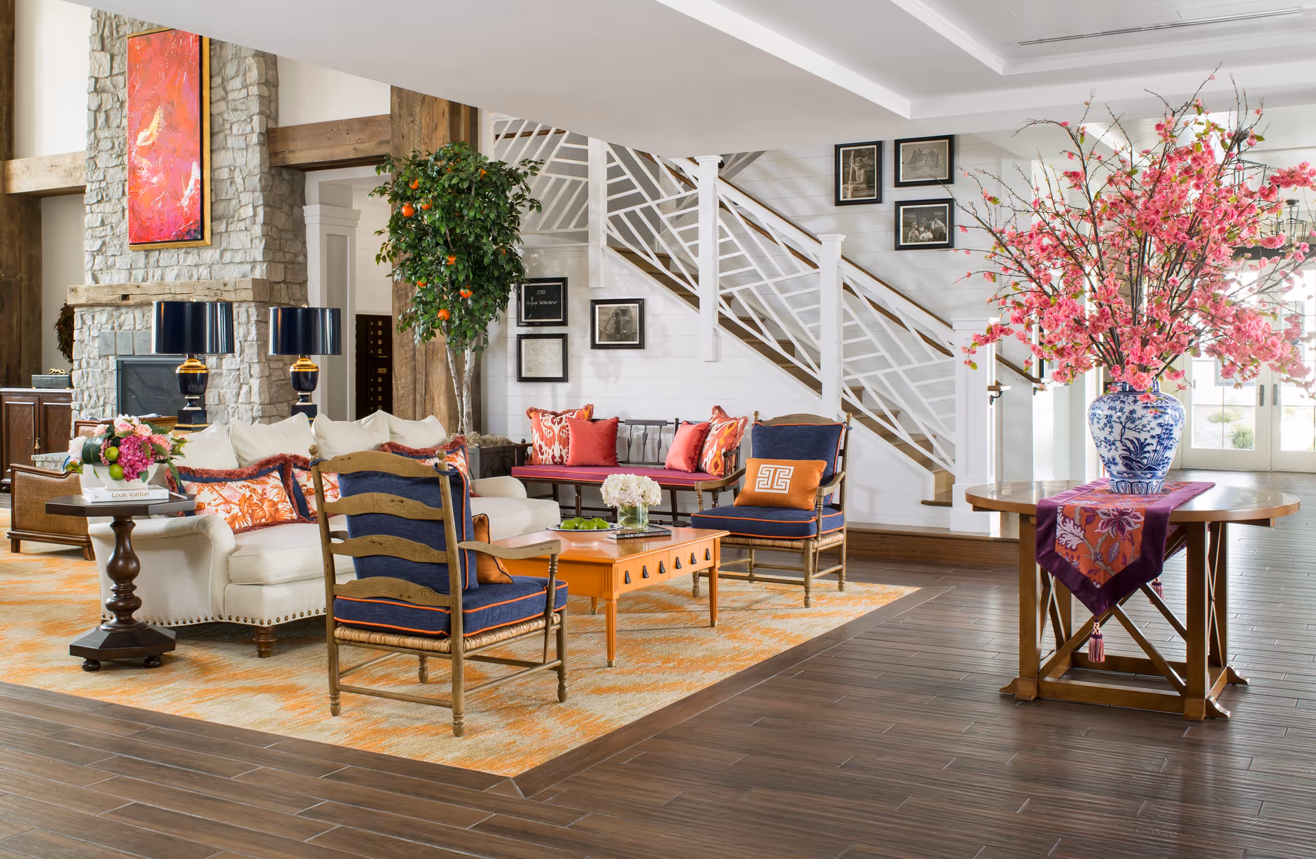 A bright and spacious living room area featuring a white sofa with colorful cushions, two blue cushioned wooden chairs, a wooden coffee table, and a side table with a flower arrangement. There is a large stone fireplace with a vibrant red and orange painting above it. A staircase with white railings and framed pictures on the wall is visible in the background. A wooden table with a large blue and white vase filled with pink flowers is positioned near the entrance with glass doors.