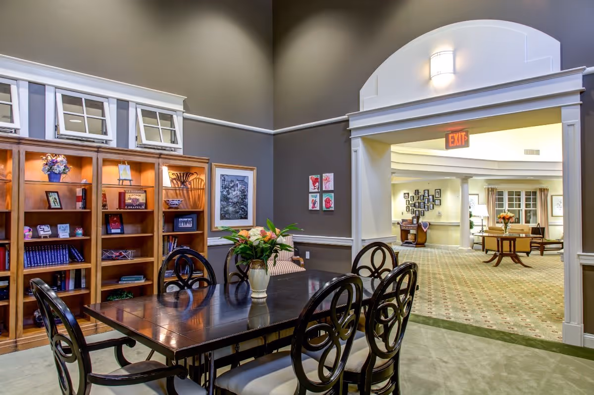 Interior view of a senior living facility showing a dining area with a dark wooden table and six chairs. A vase with flowers is placed on the table. Behind the table is a wooden bookshelf with various decorative items and books. The room has high ceilings and neutral-colored walls. An open doorway leads to a larger common area with carpeted floors, tables, chairs, and framed pictures on the walls.