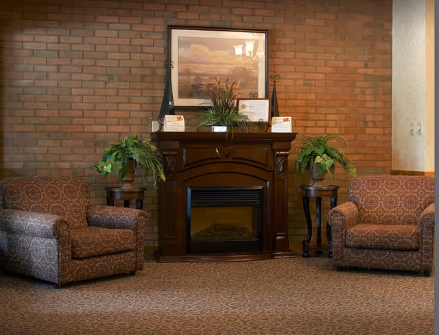 A cozy sitting area with two patterned armchairs facing a dark wooden electric fireplace. The fireplace mantel is decorated with green plants and framed certificates, and a framed landscape painting hangs on the brick wall above it. The floor is carpeted with a patterned design.