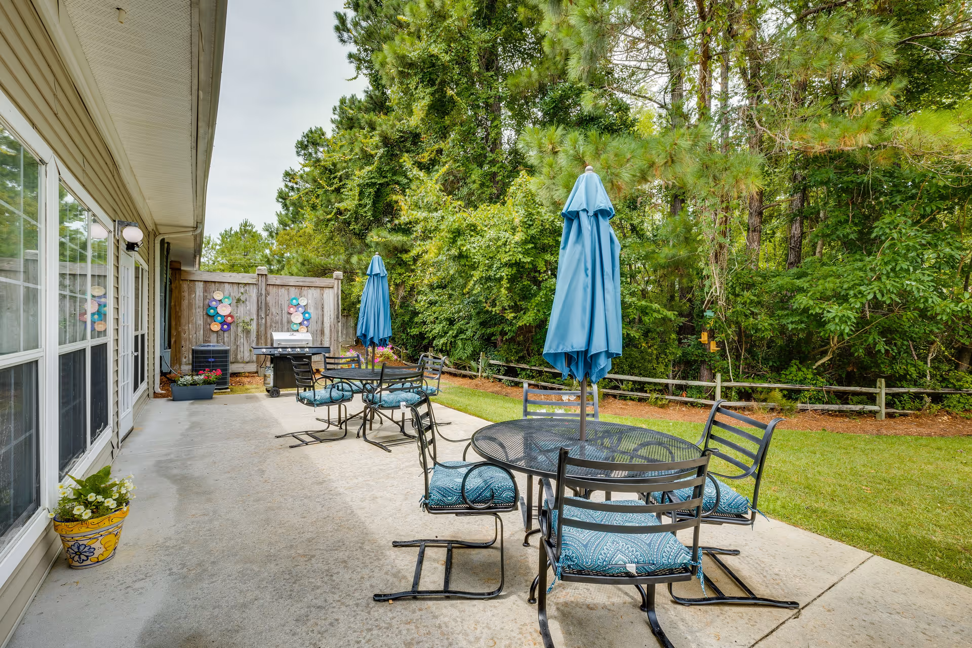 Outdoor patio area with metal tables and chairs featuring blue cushions and closed blue umbrellas. The patio is adjacent to a building with large windows and a door. There is a barbecue grill and decorative wall art on a wooden fence at the far end. The patio overlooks a grassy area bordered by trees and a wooden fence.