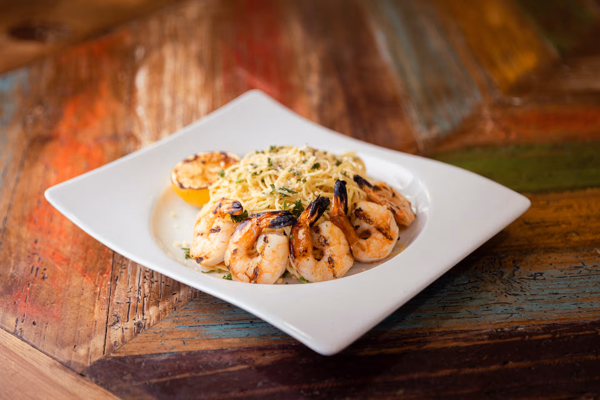 Grilled shrimp arranged around pasta with a charred lemon half on a square white plate on a rustic wooden table.