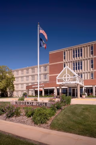 Front entrance of the Ridgeway Place senior living building with flagpoles, a stone sign, and landscaped grounds.