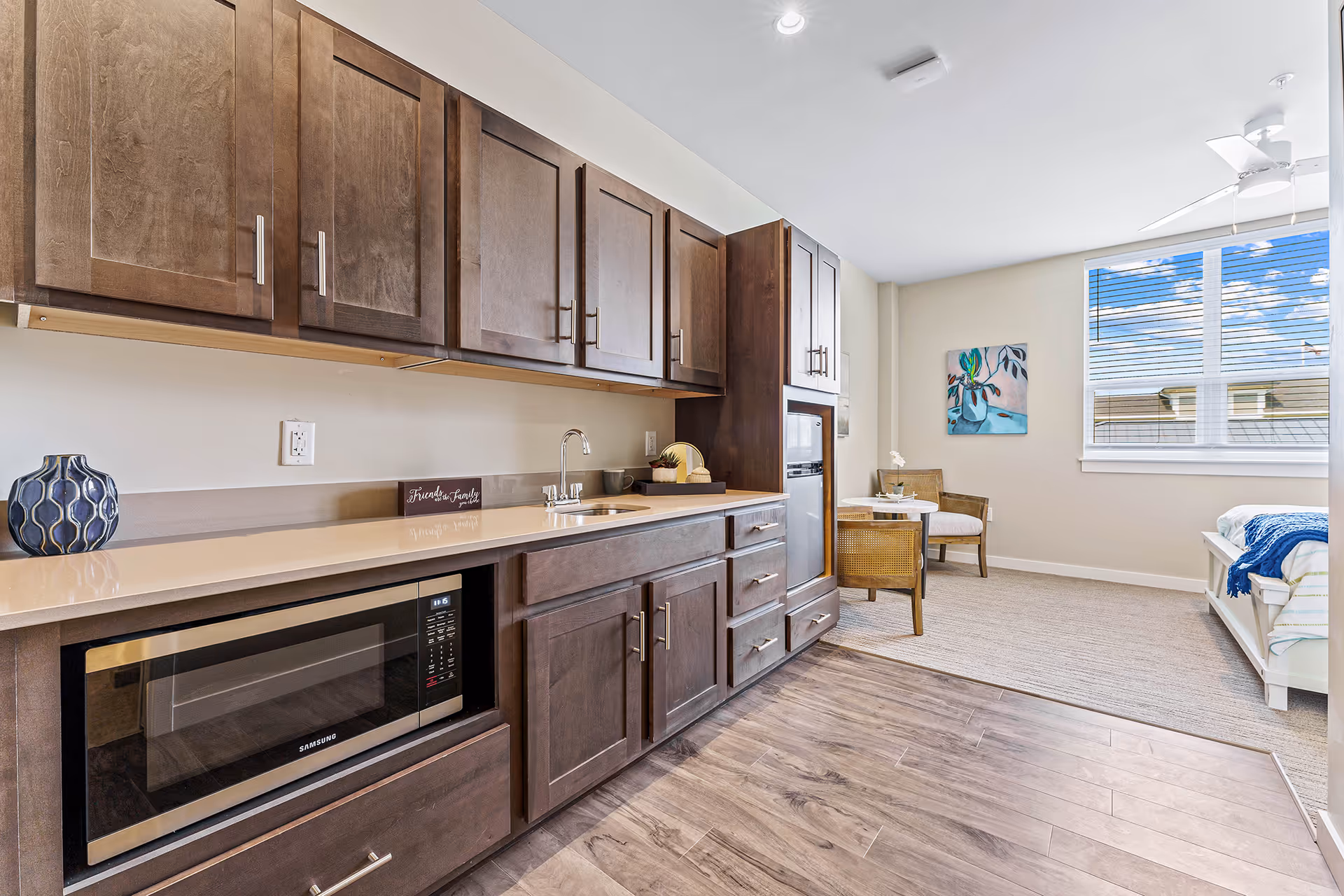 Interior view of a senior living facility room featuring a kitchenette with dark wooden cabinets, a microwave, a sink, and a countertop. Adjacent to the kitchenette is a small dining area with two chairs and a round table, a bed with a blue throw blanket, a large window with blinds, and a ceiling fan. The floor transitions from wood near the kitchenette to carpet in the living and sleeping area.