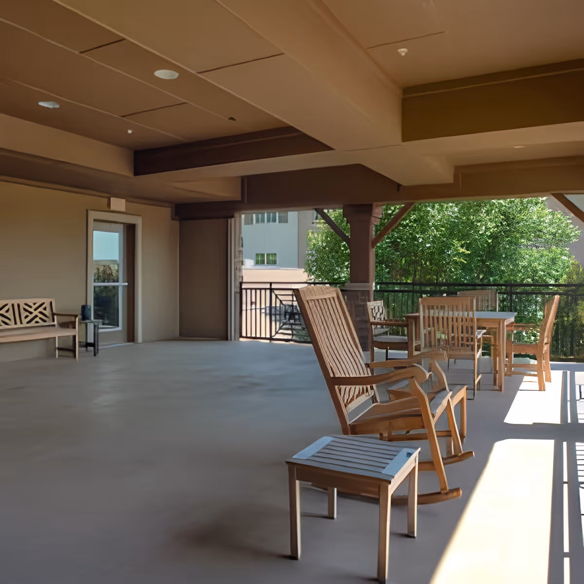 Covered outdoor patio area with wooden rocking chairs, a small wooden bench, and a table with chairs. The space is shaded with a ceiling and beams, and there is greenery visible beyond the railing.