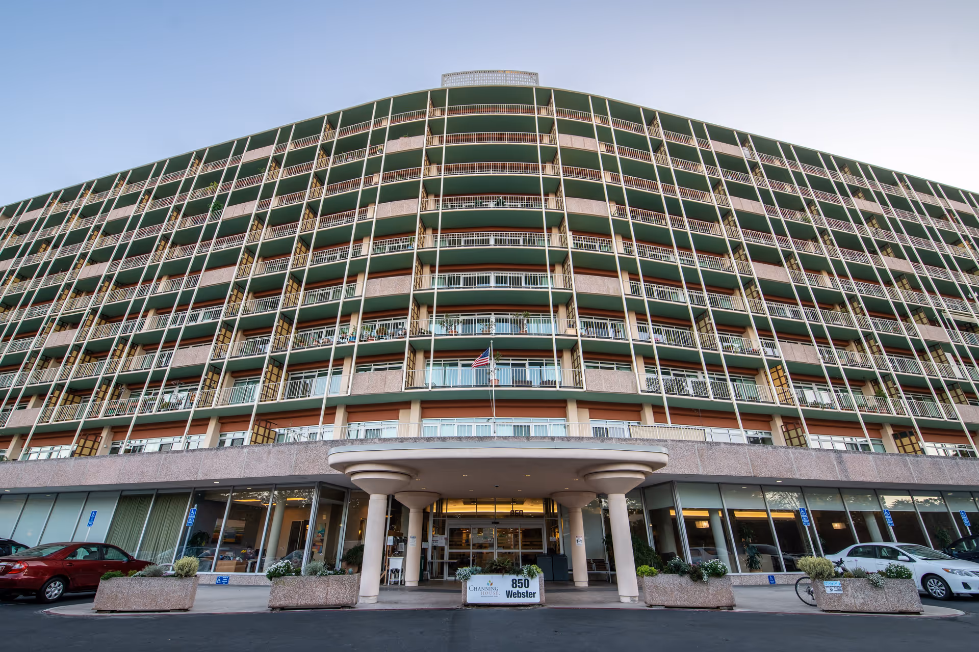 Front exterior view of Channing House, a multi-story building with balconies on each floor, an entrance canopy supported by columns, and a sign displaying the address 850 Webster. Several cars are parked in front of the building.