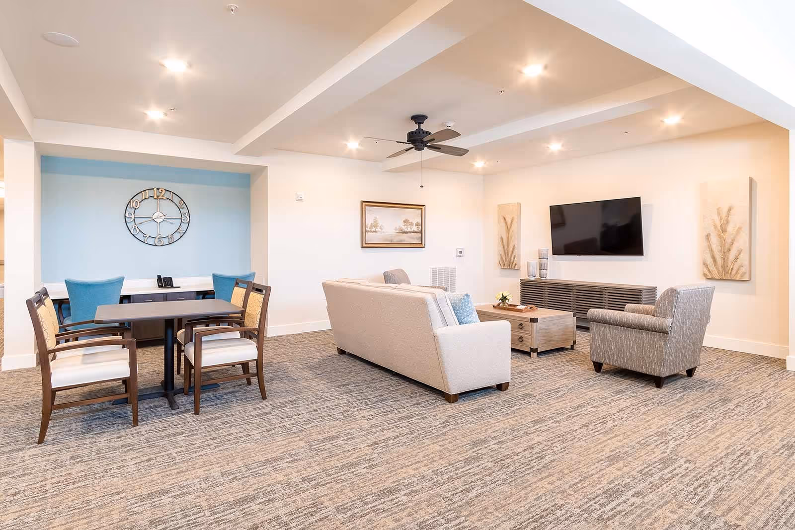 A bright and spacious living room area in a senior living facility featuring a beige sofa, a gray armchair, a wooden coffee table, a wall-mounted flat-screen TV, and decorative wall art. To the left, there is a small dining or game table with four chairs, two of which are blue and two are wooden with cushioned seats. The room has recessed lighting, a ceiling fan, and a carpeted floor.