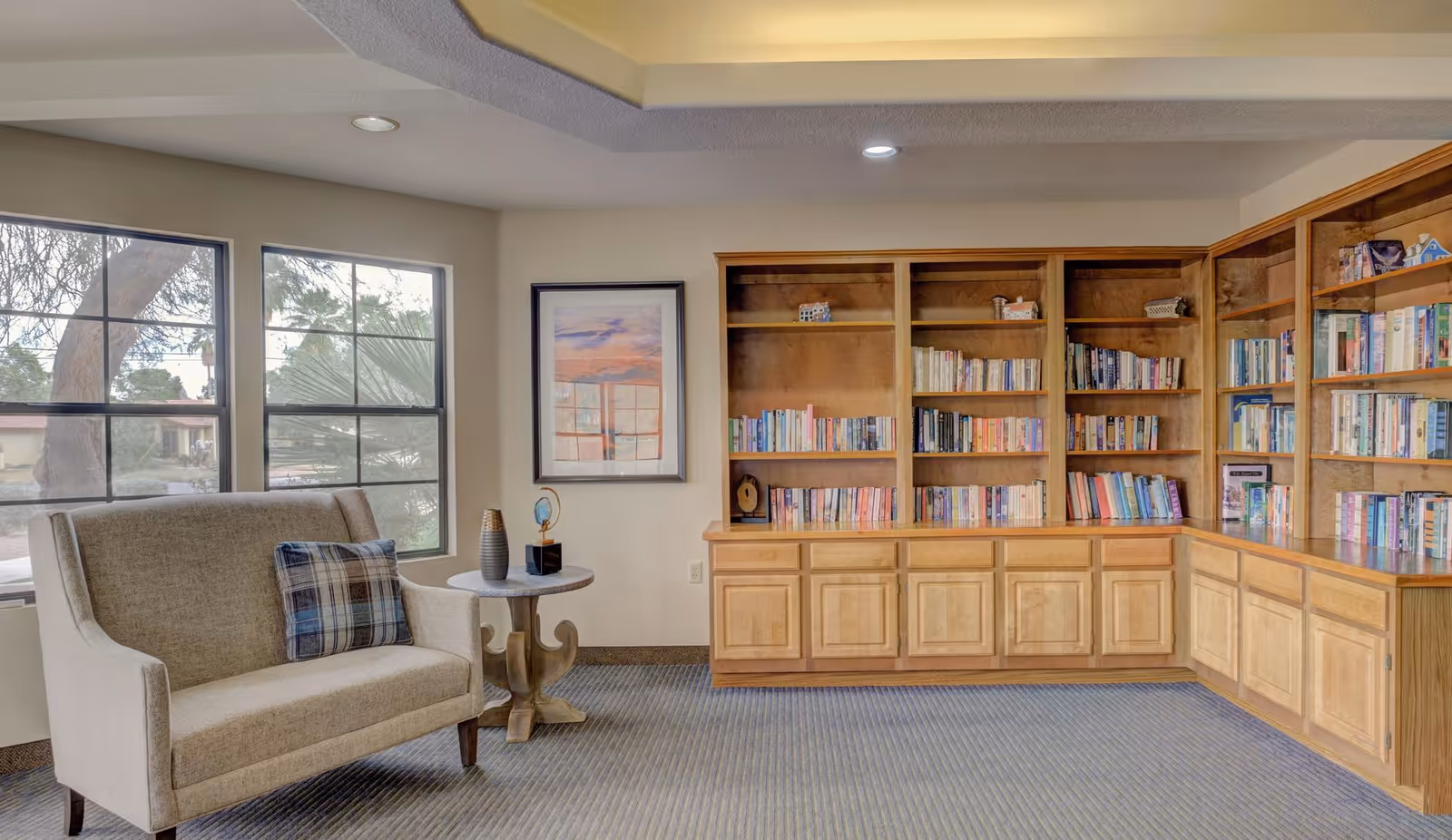 A cozy reading nook in a senior living facility featuring a beige loveseat with a plaid pillow, a small round wooden side table with decorative items, large windows letting in natural light, and built-in wooden bookshelves filled with books along the wall.