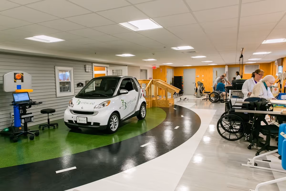 Indoor rehabilitation area with a small white car labeled Brentwood on a green and black track. Several people, including elderly individuals and caregivers, are engaged in therapy activities. There is a wooden ramp, wheelchairs, exercise equipment, and a basketball hoop attached to a computer workstation.