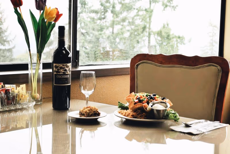 A dining table by a window set with a bottle of wine, an empty wine glass, a plate of food, a small side dish, and a vase of tulips.