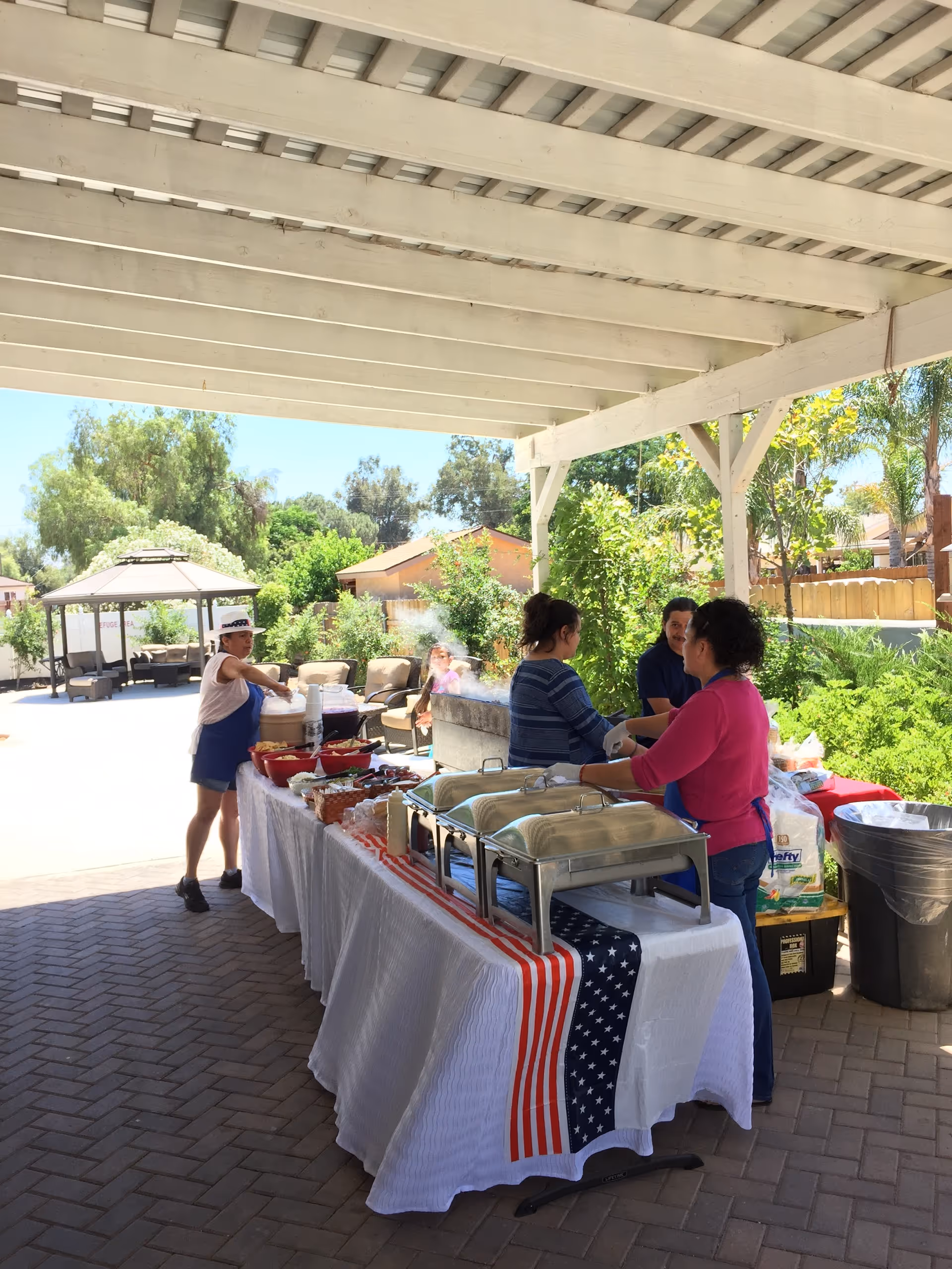 Outdoor covered patio area with a buffet table set up for serving food. Three people are behind the table preparing or serving food. The table is covered with a white tablecloth and a patriotic American flag-themed runner. There are chafing dishes, bowls, and condiments on the table. In the background, there is greenery, a gazebo with seating, and a sunny sky.