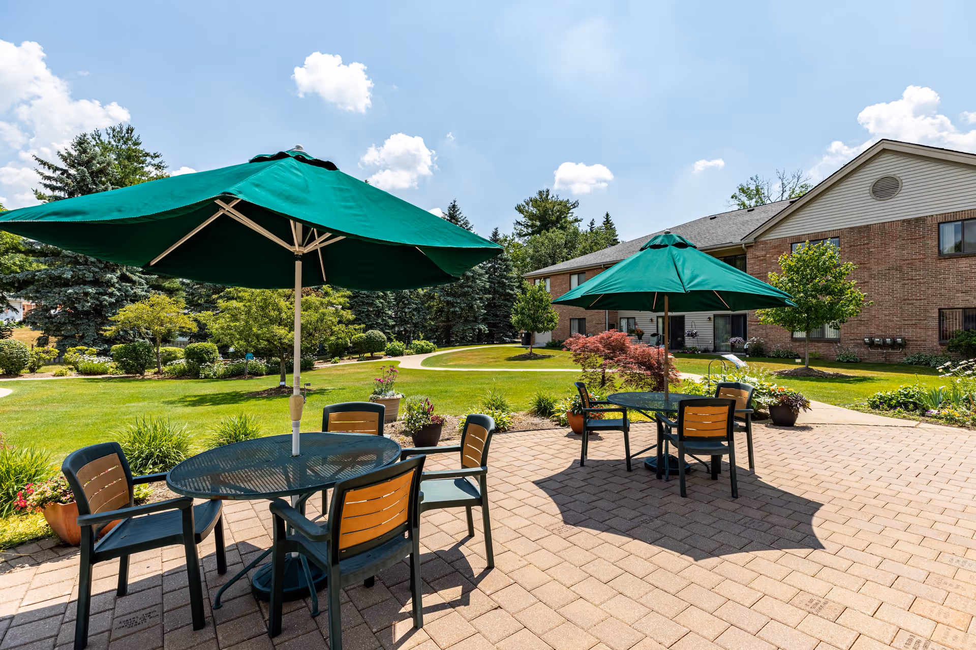 Outdoor patio area at Lourdes Senior Community with two round tables, each shaded by a large green umbrella. Each table is surrounded by four chairs. The patio is paved with bricks and overlooks a well-maintained garden with green grass, shrubs, and trees. A brick building is visible in the background under a partly cloudy blue sky.