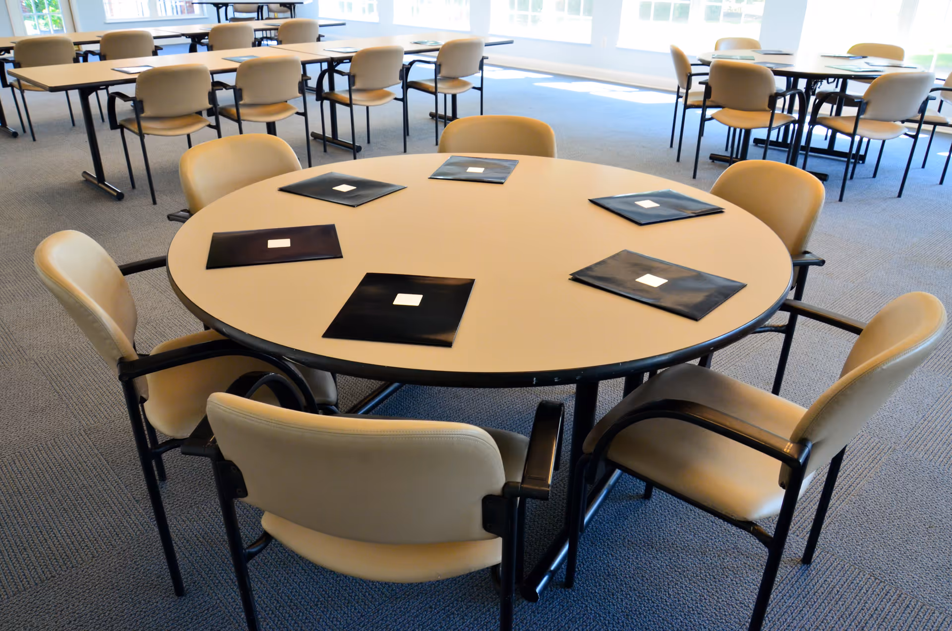 A bright dining room with multiple round and rectangular tables surrounded by beige chairs. Each round table has black folders placed on it. Large windows in the background let in natural light.