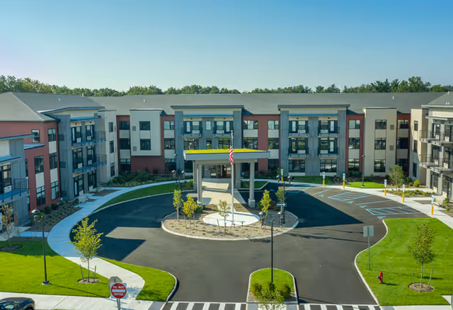 Front exterior view of a senior living facility with a circular driveway, landscaped greenery, and an American flag displayed at the entrance under a covered drop-off area. The building has multiple floors with balconies and large windows.