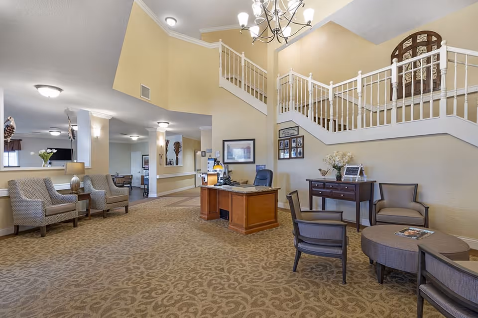 A spacious and well-lit interior common area of a senior living facility with beige walls and carpet. The area features a wooden desk with a chair, several upholstered chairs arranged around a small round table, and a side table with flowers and decorative items. A staircase with white railings leads to an upper floor, and framed pictures hang on the walls.
