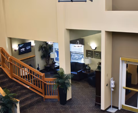 Interior view of a senior living facility lobby area with a wooden staircase, patterned carpet, potted plants, and seating areas with dark sofas and lamps. A TV is mounted on the wall above a fireplace, and there are framed pictures on the walls. The space has high ceilings and warm beige walls.