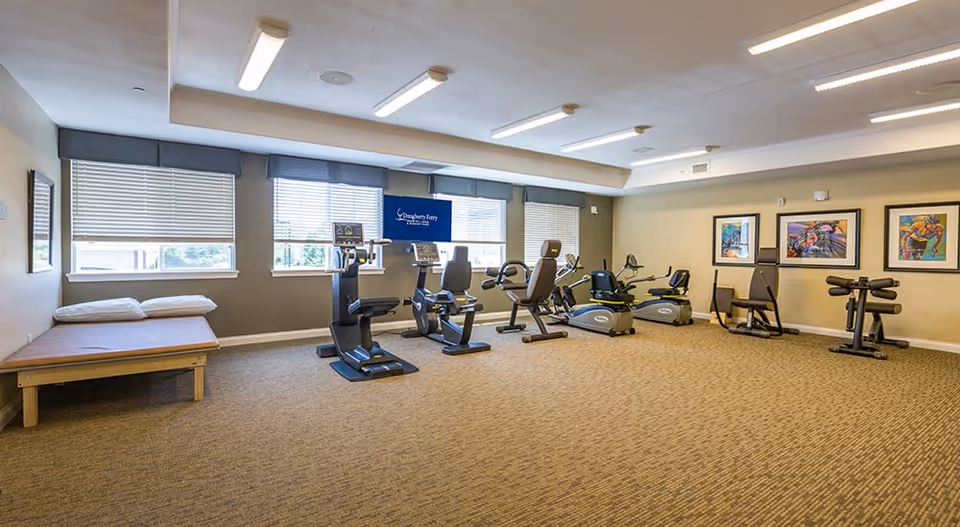 A spacious exercise room with several pieces of cardio and strength training equipment arranged along the walls. There is a wooden bench with two pillows on the left side near three windows with blinds. The walls are decorated with colorful framed artwork, and a television mounted on the wall displays the Dougherty Ferry Assisted Living and Memory Care logo.