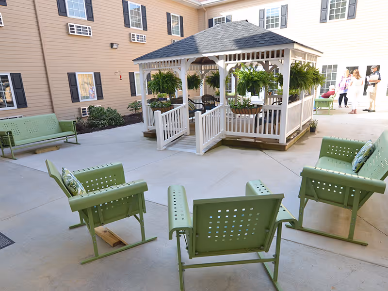 Outdoor courtyard area at a senior living facility with a white gazebo in the center decorated with hanging green plants. Surrounding the gazebo are green metal chairs and benches arranged on a concrete patio. The building exterior is beige with multiple windows and black shutters. A few people are standing and conversing in the background near the building.