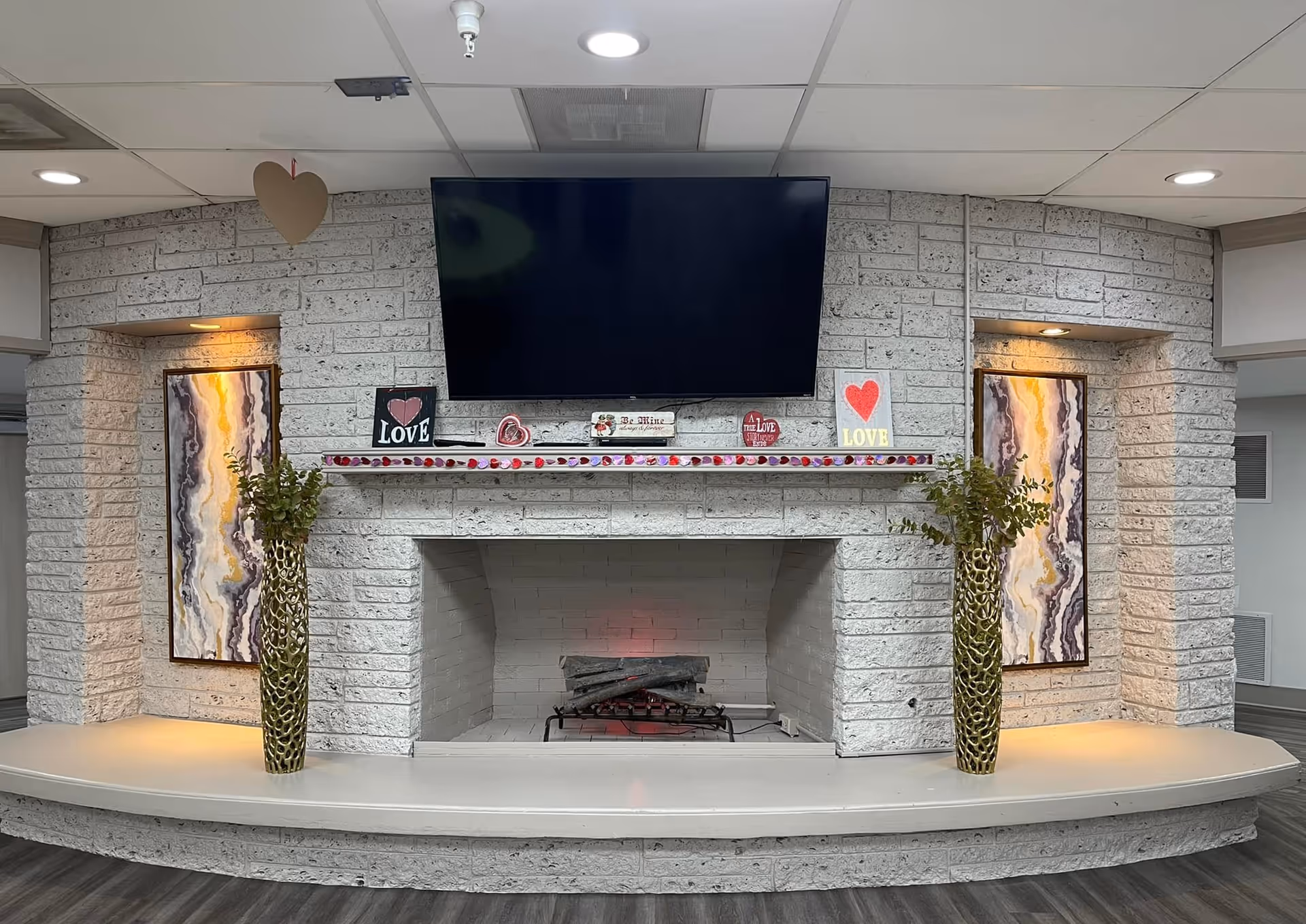 A cozy indoor fireplace with white brick surround and a mounted flat-screen TV above it. The mantel is decorated with small signs and heart-themed decorations. Two tall vases with green foliage are placed symmetrically on either side of the fireplace. The wall features two illuminated abstract paintings in recessed niches, and the floor is dark wood.