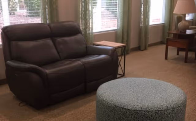 A cozy living room area featuring a dark brown leather loveseat, a round textured ottoman in front, a small wooden side table with a metal frame, and a wooden end table with a lamp. The room has large windows with green patterned curtains allowing natural light to enter.
