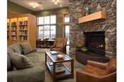 Cozy common lounge with upholstered seating, a stone fireplace, bookshelves, and a large window.