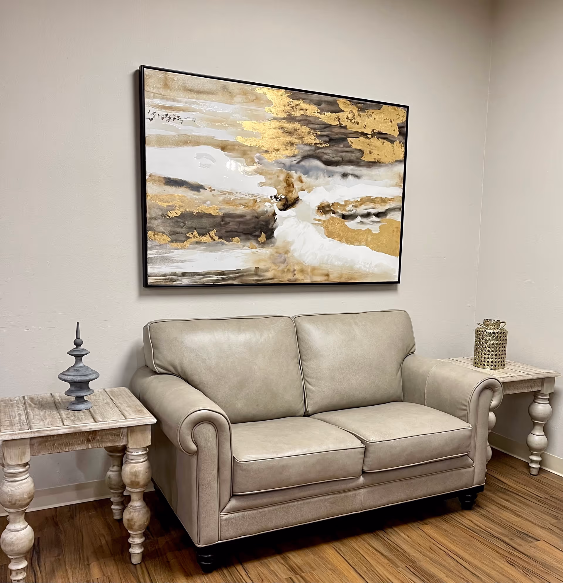 A beige leather loveseat sofa is positioned against a light-colored wall with a large abstract painting featuring gold, white, and gray tones hanging above it. On either side of the sofa are matching wooden side tables with decorative items placed on top. The floor is wooden with a warm finish.