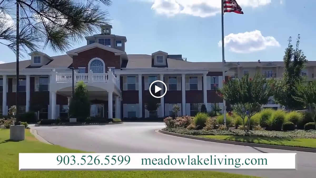 Front exterior view of Meadow Lake Senior Living facility with a driveway, landscaped garden, American flag on a flagpole, and a clear blue sky with some clouds.