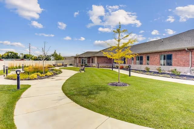 A sunny outdoor garden area at Lakewood Memory Care & Assisted Living featuring a curved concrete walkway, green grass, small trees, landscaped flower beds, benches, and a red brick building with multiple windows under a blue sky with scattered clouds.