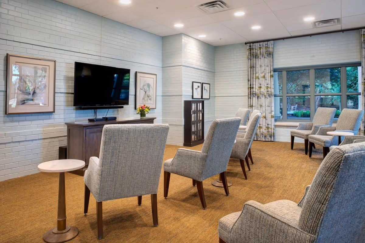 A cozy common area with several upholstered armchairs arranged in rows facing a wall-mounted flat-screen TV. The room has light blue painted brick walls adorned with framed artwork, a wooden cabinet beneath the TV, a small round side table, and a large window with patterned curtains letting in natural light and showing greenery outside.