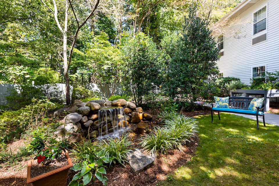 A peaceful outdoor garden area with a small rock waterfall surrounded by green plants and trees. There is a black metal bench with blue cushions and patterned pillows on a grassy lawn next to a white building with multiple windows.