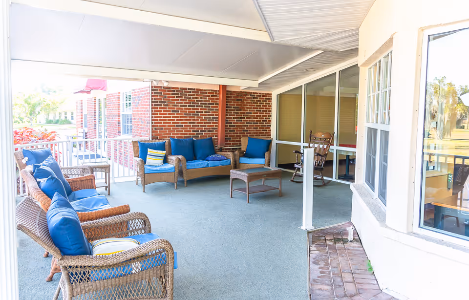 Covered outdoor patio area with wicker seating including chairs and a loveseat with blue cushions, a small wicker coffee table, and a wooden rocking chair. The patio has a brick wall on one side and large windows on another, overlooking a green outdoor space.