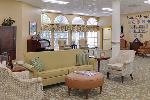 A well-lit living room area with a beige sofa, two patterned armchairs, a round brown ottoman, and side tables. Large windows with arched tops and beige valances let in natural light. In the background, there is a 'Wall of Honor' display with plaques and an American flag. The room has a patterned carpet and neutral-colored walls.