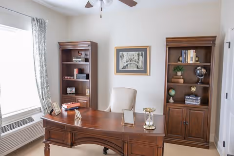 A well-lit office or study room with a large wooden desk in the center, an office chair behind it, two wooden bookshelves filled with books and decorative items, a framed picture on the wall, and a window with patterned curtains letting in natural light.
