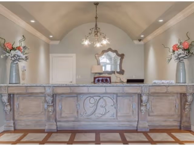 Reception desk area with a large wooden counter featuring ornate carvings and the initials 'PB' in the center. Behind the desk is a wall with a decorative mirror, a lamp, and a small chair. Two large vases with floral arrangements are placed symmetrically on each end of the counter. The ceiling has recessed lighting and a chandelier hanging above the desk.