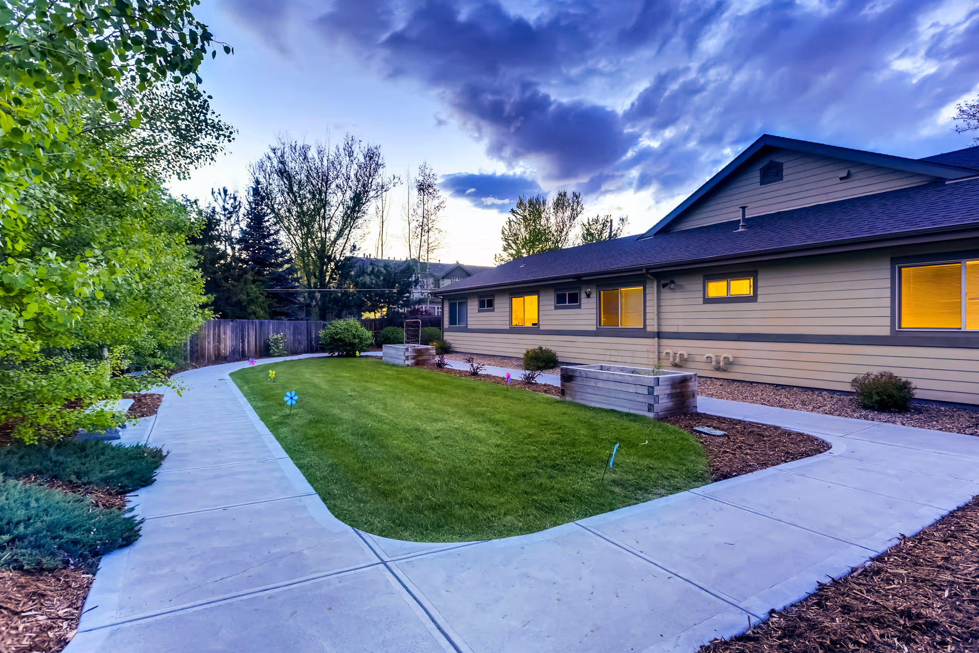 Exterior view of a single-story assisted living building at dusk with lit windows, a curved concrete walkway, and a grassy landscaped courtyard.