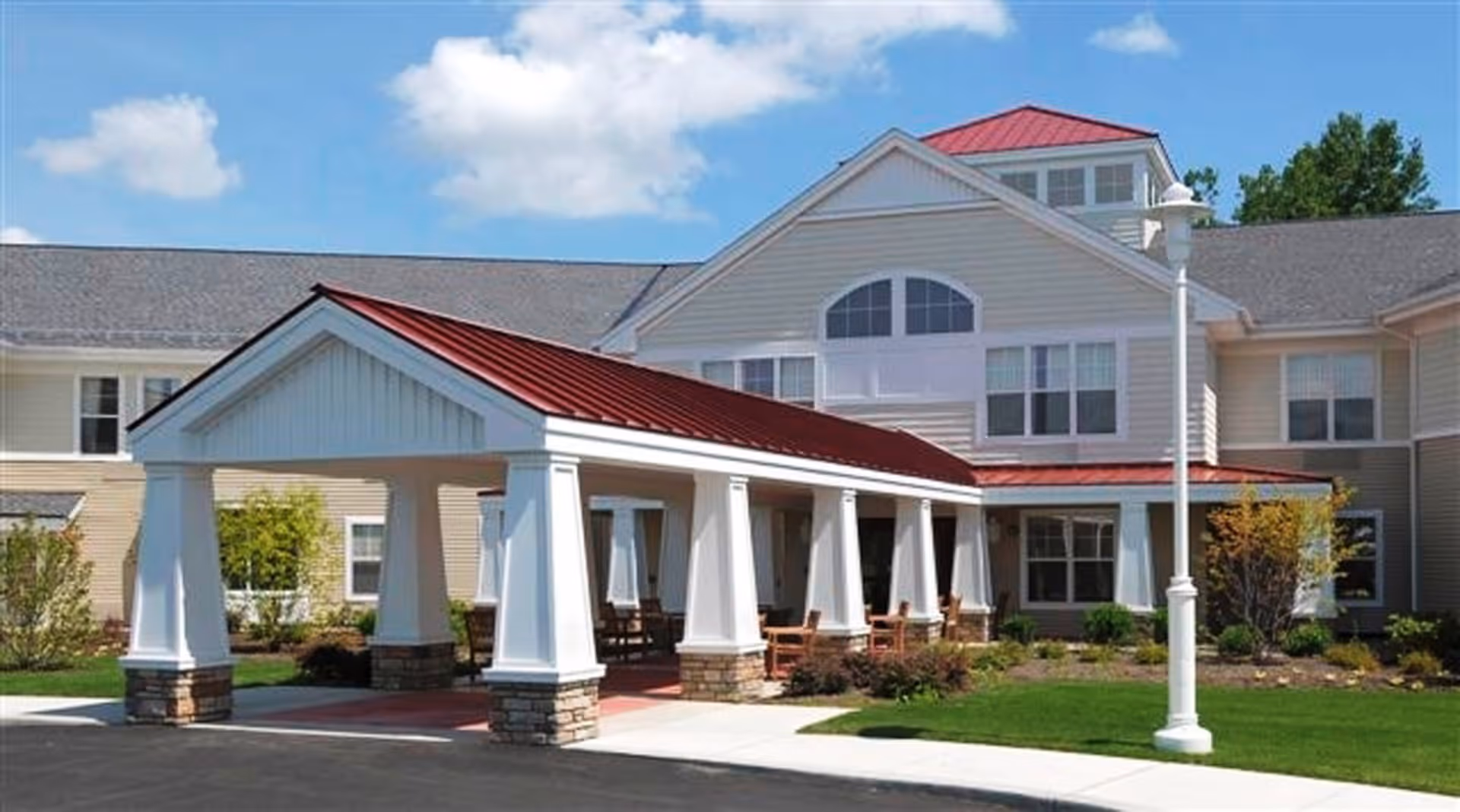 Covered entrance with white columns and a red metal roof in front of a beige multi-story senior living building.