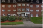 Front exterior view of a multi-story brick building with multiple windows and a central entrance accessed by stairs with railings. There are bushes and a lawn in front of the building.