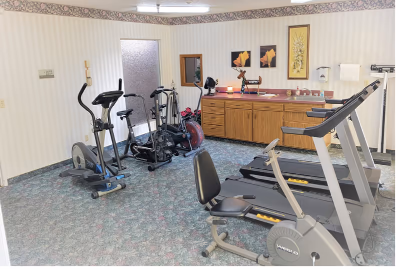 A small exercise room with various fitness equipment including a treadmill, stationary bike, elliptical machine, and rowing machine. The room has patterned carpet, wallpaper with a floral border near the ceiling, a wooden cabinet with a countertop, and framed floral artwork on the wall.