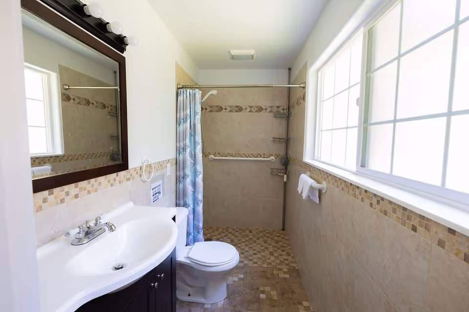 A bright bathroom with a white sink and dark wood vanity, a large mirror with lights above it, a toilet, and a walk-in shower with a blue patterned shower curtain. The walls and floor are tiled with beige and brown mosaic accents, and there is a large window letting in natural light.