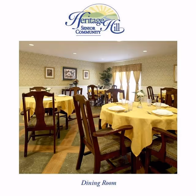 Dining room in Heritage Hill Senior Community with round tables covered in yellow tablecloths, set with plates, glasses, and small flower vases. Wooden chairs surround the tables, and the room has patterned wallpaper, framed artwork, and large windows with curtains letting in natural light.