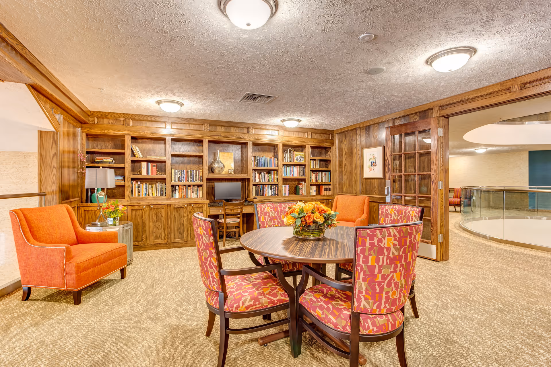 A cozy interior room with wood-paneled walls and built-in bookshelves filled with books and decorative items. There is a round wooden table with four colorful patterned chairs around it, and a floral centerpiece on the table. Two orange armchairs are placed near the bookshelves. The room has a beige carpet and ceiling lights, with a glass-paneled wooden door leading to a hallway with a curved railing.