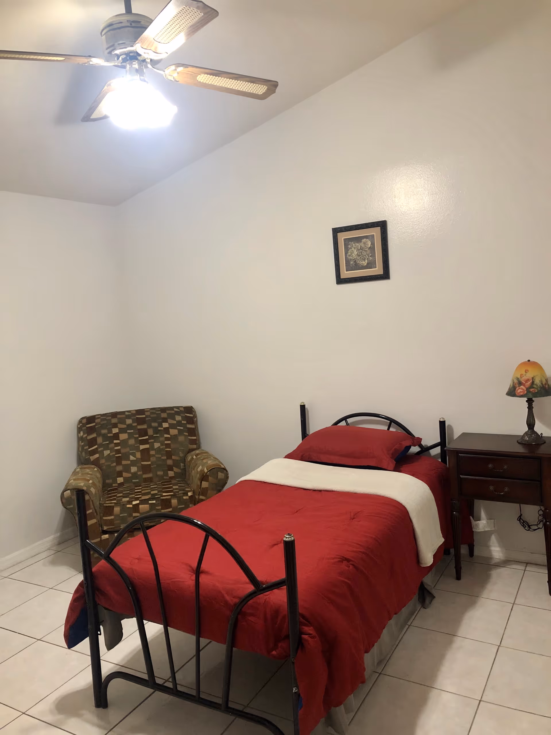 Small bedroom with a single metal bed dressed in red bedding, a wooden nightstand with a lamp, a patterned armchair, a ceiling fan, and a framed picture on a white wall.