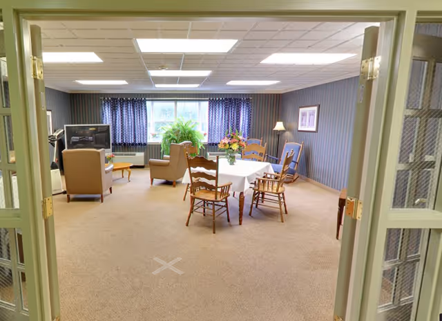 A cozy senior living common area with beige carpet, a table covered with a white tablecloth and surrounded by wooden chairs, two armchairs facing a television, a large green plant near the window with blue patterned curtains, and a floor lamp in the corner. The room is viewed through open double doors with glass panels.