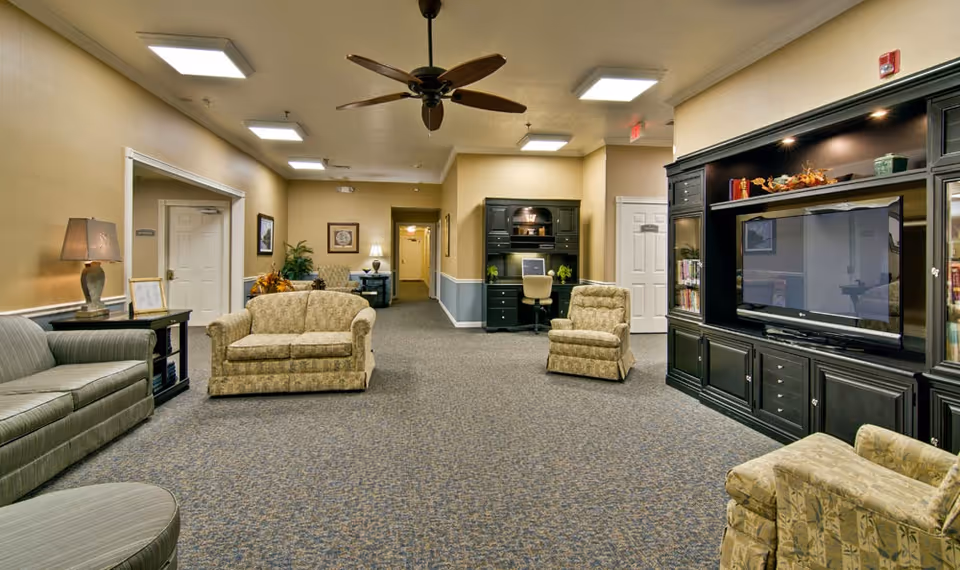 A spacious senior living facility common area with multiple upholstered armchairs and sofas arranged around a large black entertainment center with a flat-screen TV. The room has beige walls with a blue wainscot, carpeted floor, ceiling fans, and recessed lighting. There is a desk with a chair and plants in the background, and doors leading to other rooms.