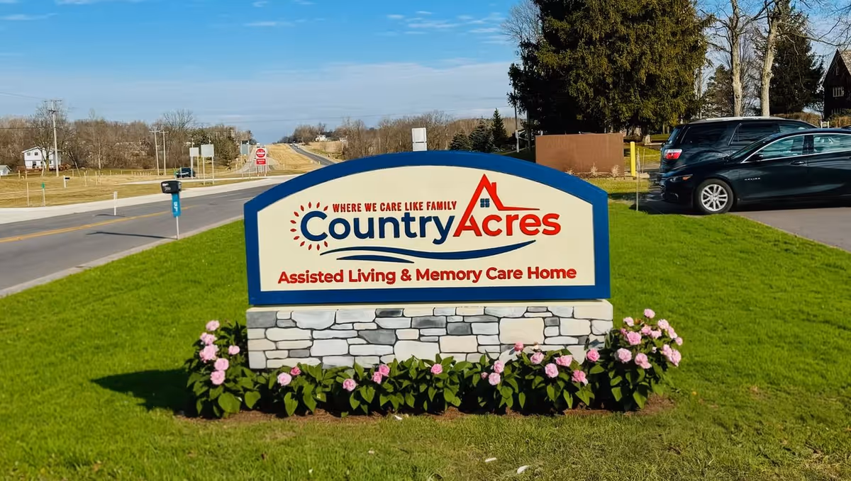 Outdoor view of a sign for Country Acres Assisted Living & Memory Care Home, placed on a grassy area with pink flowers in front. The sign has a blue border and stone base, with a road and parked cars visible in the background under a clear sky.