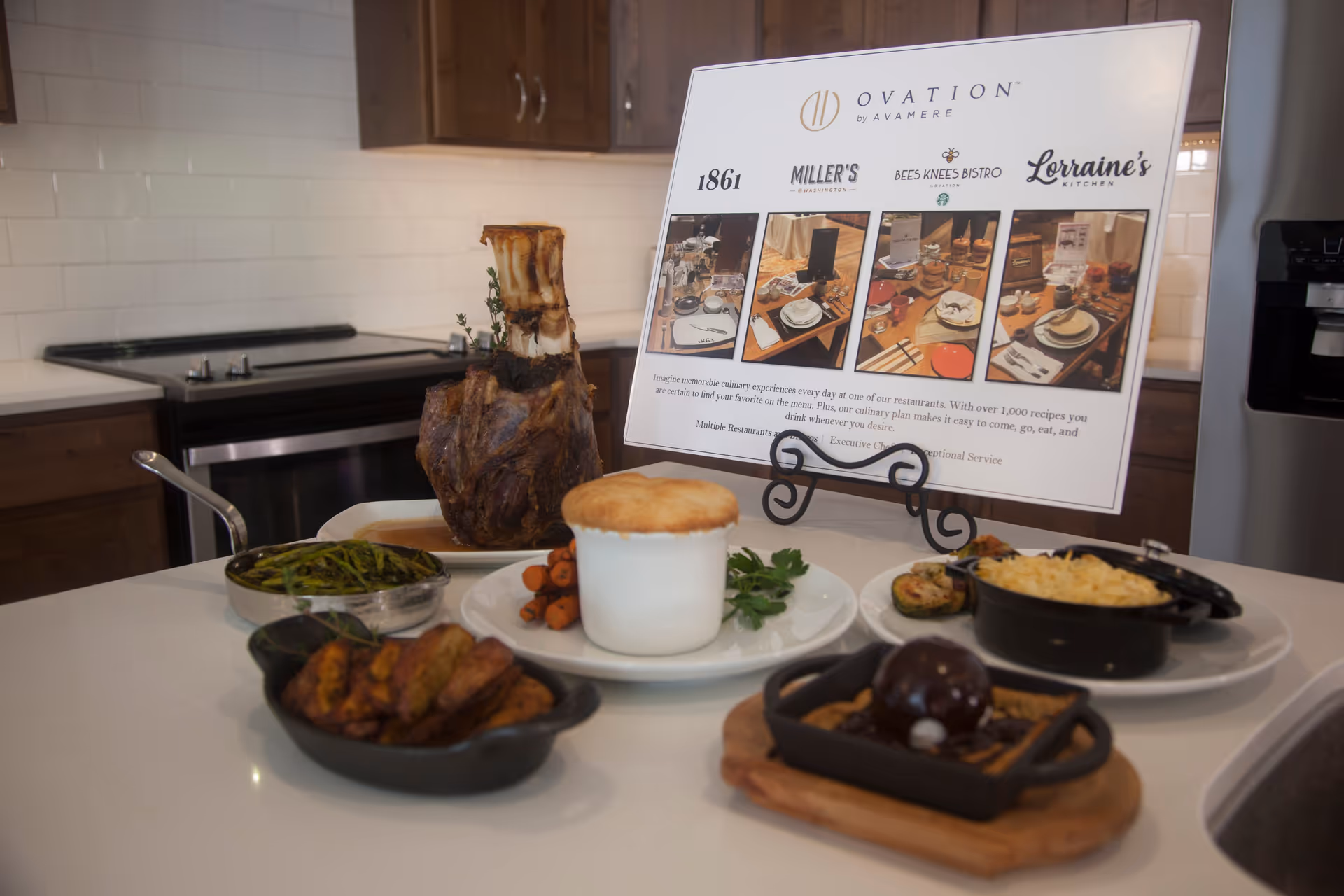 A kitchen countertop displaying a variety of cooked dishes including a large roasted bone-in meat, sautéed green beans, roasted carrots, a pot pie, macaroni and cheese, and a dessert with a chocolate sphere. Behind the food is a sign on a stand featuring the Ovation by Avamere logo and information about multiple restaurants within the facility.
