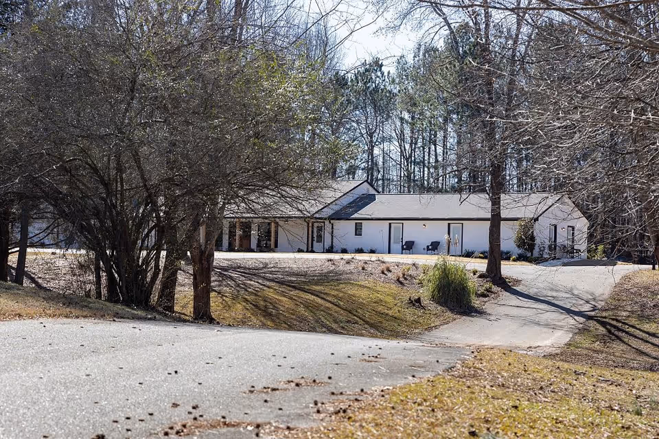 A single-story white building with a gray roof surrounded by leafless trees and a curved driveway leading up to it on a clear day.