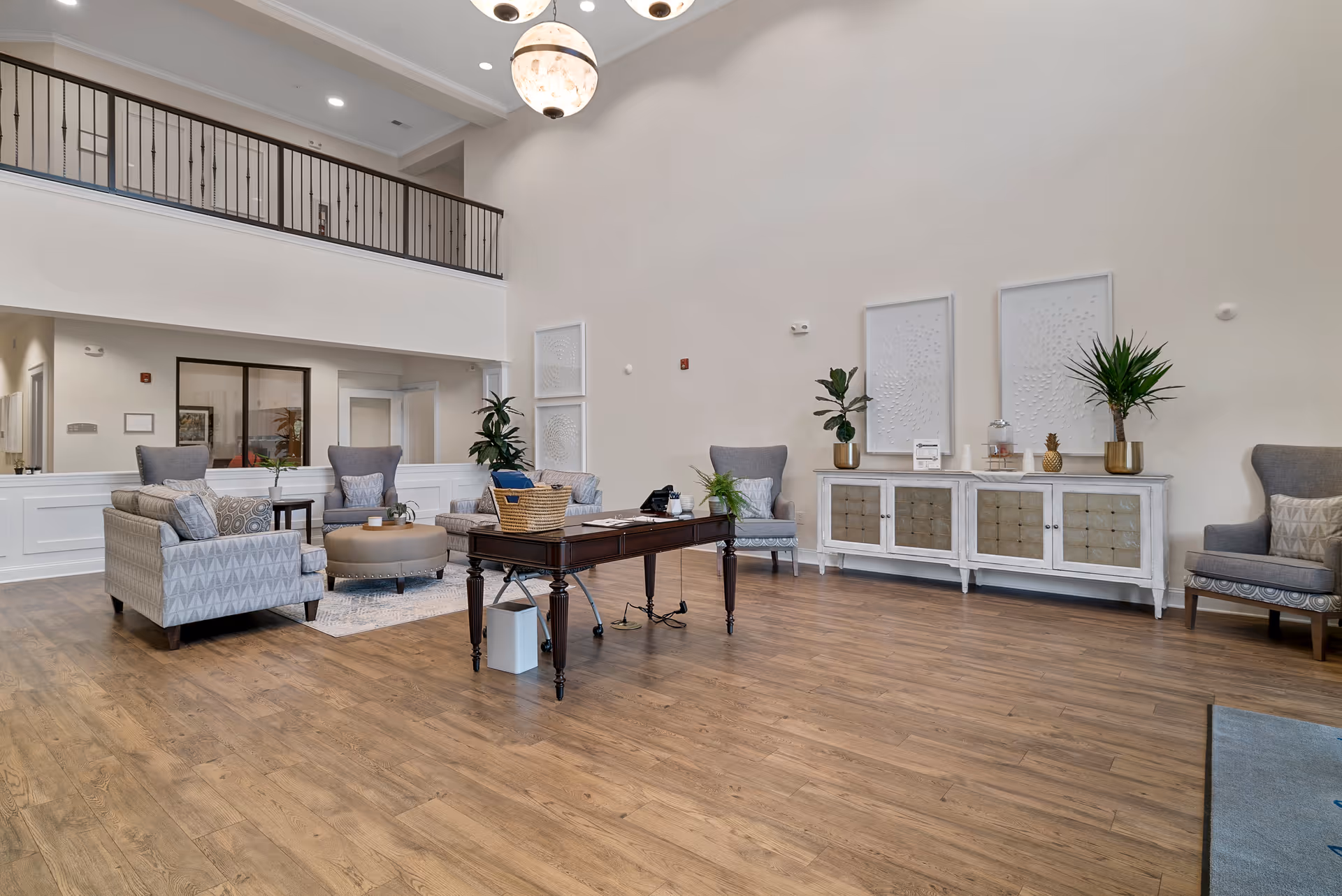 Spacious and well-lit living room area with high ceilings, featuring a wooden floor, a dark wooden table with a basket and phone on it, several upholstered armchairs and a round ottoman arranged around a small rug. There is a white sideboard against the wall with decorative plants and framed artwork above it. A balcony railing is visible on the upper level.