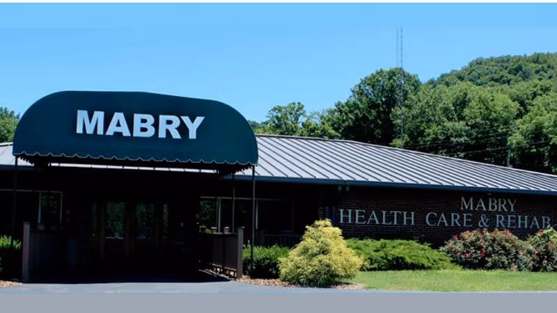 Exterior front view of Mabry Health Care & Rehab Center building with a green awning displaying the word 'MABRY' and a brick facade with the facility name on the wall, surrounded by greenery and trees under a clear blue sky.