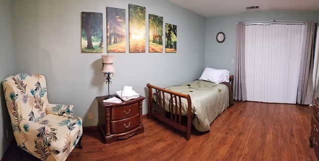 A simple bedroom with a single wooden bed covered with a green bedspread, a wooden nightstand with a lamp and folded towels, a patterned armchair, a wall clock, vertical blinds covering a sliding glass door, and a multi-panel wall art depicting a forest scene.